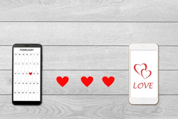 Valentine day concept, on white wooden background isolated red paper hearts and two smartphones, calendar and greeting on smartphone screen — Stockfoto
