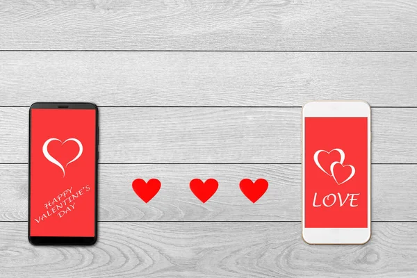 Valentine day concept, on white wooden background isolated red paper hearts and two smartphones, greeting on smartphone screen — Stockfoto