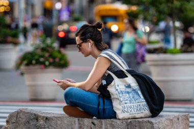 New York, Usa - 21 Haziran 2019: Caddenin kenarında bir kadın bir taşın üzerine oturur ve arka planda bulanık şehir manzarası olan akıllı bir telefondan müzik dinler.