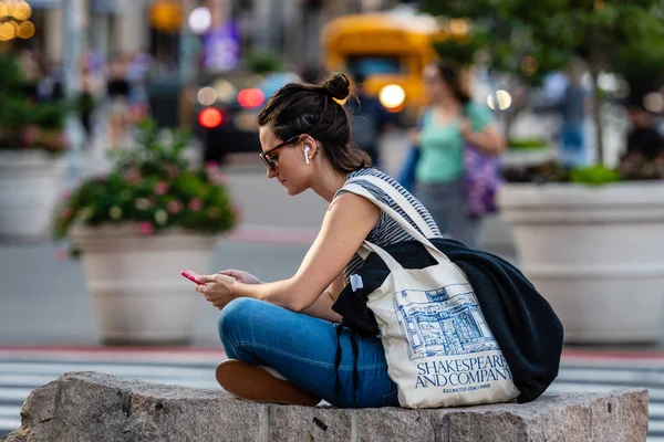 Nueva York, Estados Unidos - 21 de junio de 2019: al borde de la calle, una mujer se sienta en una piedra y escucha música desde un teléfono inteligente, paisaje urbano borroso en el fondo —  Fotos de Stock