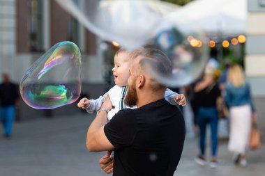 Old Town Square, Riga, Latvia - August 16, 2019: Bearded man with a child on his hands watches and rejoices at the gigantic soap bubbles