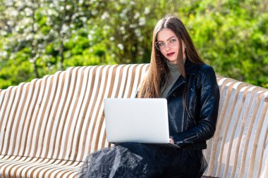 Gözlüklü genç bir kadın parktaki bankta dizüstü bilgisayarla oturuyor güneşli ve rüzgarlı bir bahar gününde kameraya bakıyor ev ofisinden uzakta herhangi bir yerde çalışıyor.