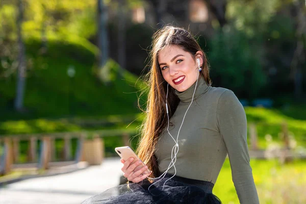 young smiling woman sit on bench in park outdoors listening music with headphones using mobile phone on a sunny and windy day, the concept of leisure in outdoor