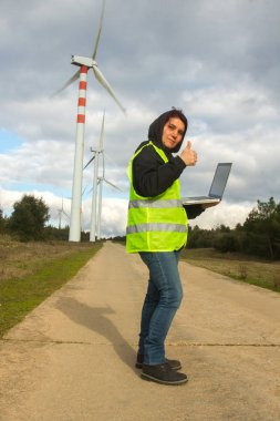 Elinde bilgisayar ve sarı yansıtıcı ceketi olan mühendis kadın rüzgar türbininin altında gülümsüyor.