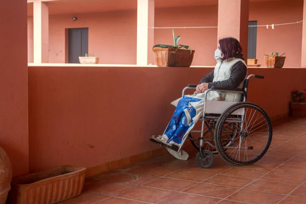 disabled woman alone in a wheelchair with a protective mask looking out from the balcony of the house staying home for the quarantine