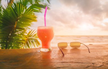 Bir bardak portakal suyu ve deniz günbatımı gökyüzünde ahşap bir masa koymak güneş gözlüğü.