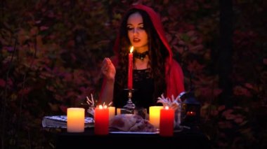 Kırmızı pelerin içinde çekici güzel esmer cadı mistik sonbahar ormanda çağrıştırıyor. büyücülük bir kalp, tavuk budu ve kirpi iğne ile