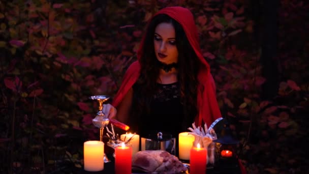 Atractiva bruja morena preciosa con libro en capa roja evoca en el bosque místico de otoño. brujería con corazón, patas de pollo y agujas de puercoespín — Vídeo de stock