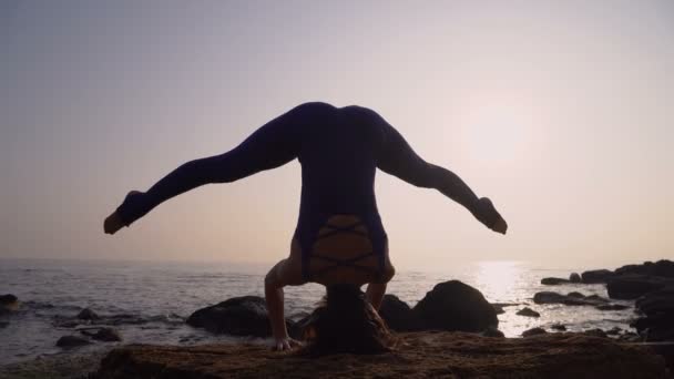 Молодая женщина в купальнике практикует йогу на пляже над морем на удивительном восходе солнца. Озил, спорт, йога и концепция здорового образа жизни. Девушка стоит на голове и бечевке — стоковое видео
