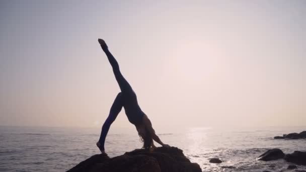 Jovem mulher em bodysuit praticando ioga na praia acima do mar ao nascer do sol incrível. Fitness, esporte, ioga e conceito de estilo de vida saudável. Menina fazendo um suporte de perna e cordel — Vídeo de Stock