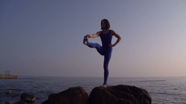 Mujer joven en traje de cuerpo practicando yoga en la playa sobre el mar al amanecer increíble. Fitness, deporte, yoga y estilo de vida saludable. Chica haciendo una pierna de pie y cordel — Vídeos de Stock