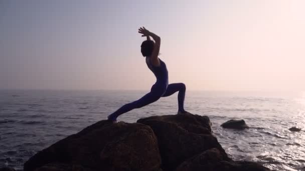 Jovem mulher em bodysuit praticando ioga na praia acima do mar ao nascer do sol incrível. Fitness, esporte, ioga e conceito de estilo de vida saudável. Menina fazendo complicado ioga asana — Vídeo de Stock