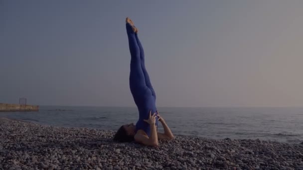 Mujer joven en traje de cuerpo practicando yoga en la playa sobre el mar al amanecer increíble. Fitness, deporte, yoga y estilo de vida saludable. Chica haciendo yoga complicado asana — Vídeo de stock