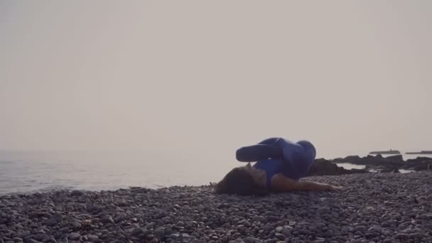 Молодая женщина в купальнике практикует йогу на пляже над морем на удивительном восходе солнца. Озил, спорт, йога и концепция здорового образа жизни. Девушка делает сложную йогу асана — стоковое видео