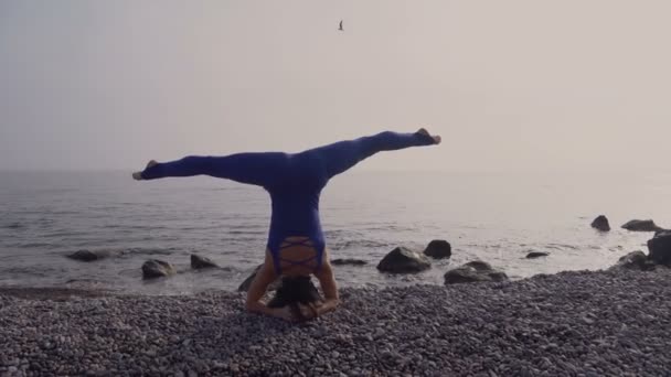 Mujer joven en traje de cuerpo practicando yoga en la playa sobre el mar al amanecer increíble. Fitness, deporte, yoga y estilo de vida saludable. Chica haciendo de pie en la cabeza y el cordel — Vídeo de stock