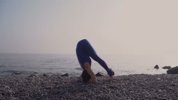 Young woman in bodysuit practicing yoga on the beach above sea at amazing sunrise. Fitness, sport, yoga and healthy lifestyle concept. Girl doing stand on head and twine — Stock Video