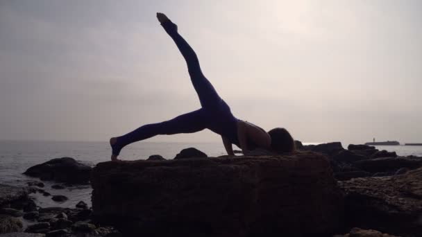 Молодая женщина в купальнике практикует йогу на пляже над морем на удивительном восходе солнца. Озил, спорт, йога и концепция здорового образа жизни. Девушка стоит на голове и бечевке — стоковое видео