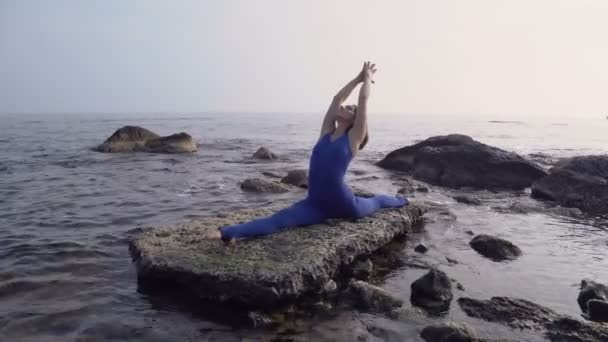 Jonge vrouw in Romper beoefenen van yoga op het strand boven zee bij fantastische zonsopgang. Fitness, sport, yoga en gezonde levensstijl concept. Meisje doet ingewikkelde yoga asana — Stockvideo