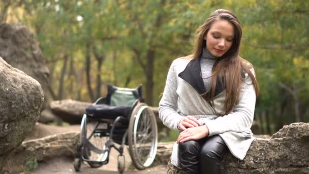 Приваблива дівчина на інвалідному кріслі, що сидить на камені в осінньому парку. Інвалідність дозвілля — стокове відео
