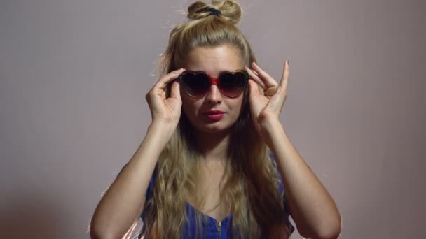Güzel seksi sarışın kız mavi elbise aşk şekil güneş gözlüğü stüdyoda ışık arka plan ile poz içinde. — Stok video