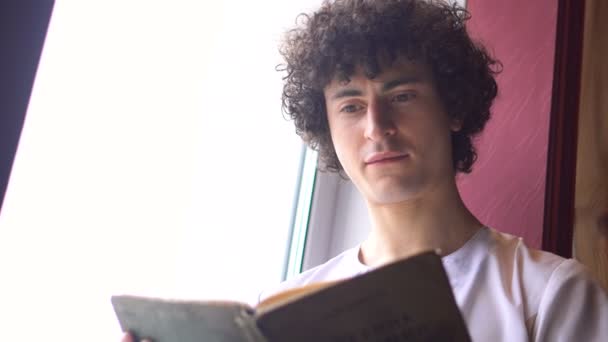 Joven hombre rizado en paño blanco está sentado en la ventana de la enfermedad y lectura del libro con la luz del día de la ventana — Vídeos de Stock