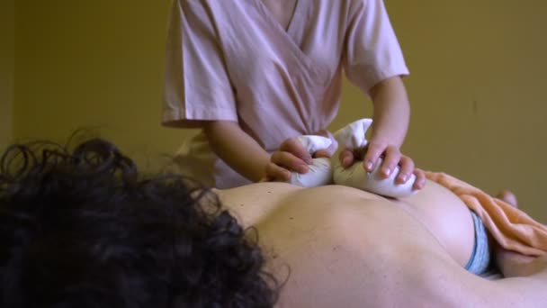 Массажистка делает массаж теплыми тряпичными пакетами молодому человеку — стоковое видео