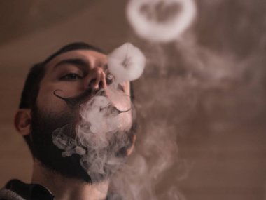 Sakal ve Mustages Vaping elektronik sigara olan adam. Vaper Hipster duman Buharlaştırıcı ve Exhals duman halkaları.