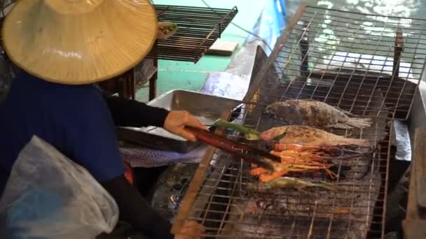 Una mujer tailandesa en paño tradicional cocinando camarones grandes en un fuego de barbacoa en el barco en Floating Market, Bangkok . — Vídeo de stock