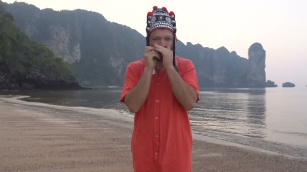 Freaky Caucasian Man in Traditional Thai Womens Hat Playing on his Harp at the Morning Twilight on the Coastline of Beautiful Tropical Rocky Cliff Lagoon in Krabi, Tailandia . — Vídeo de stock