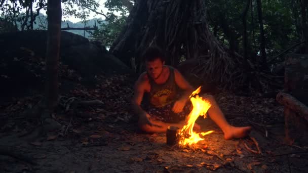 Uomo selvaggio caucasico che brucia piccolo falò nella foresta tropicale al crepuscolo per bollire il bollitore con riso per cena . — Video Stock