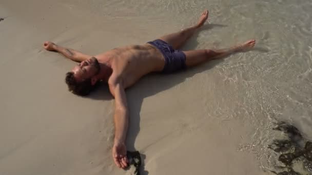 Χαλαρωτικό άντρας ξαπλωμένος στην παραλία λευκή άμμο, πλένονται με νερό ζεστό Azure στον ωκεανό. — Αρχείο Βίντεο