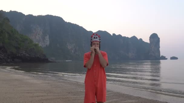 Freaky kaukázusi Férfi hagyományos Thai női kalap játszik a hárfa a reggel Twilight, a partvidék, gyönyörű trópusi sziklás Cliff lagúna-Krabi, Thaiföld. — Stock videók