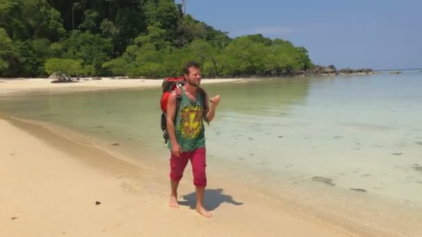 Glücklicher Wanderer mit Touristenrucksack, der auf seinem Shaker spielt, während er an der Küste der warmen, azurblauen tropischen Lagune spaziert — Stockvideo