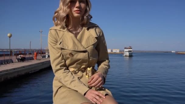 Atractiva joven rubia en trinchera con maleta vintage está sentada en el muelle Jacht — Vídeos de Stock