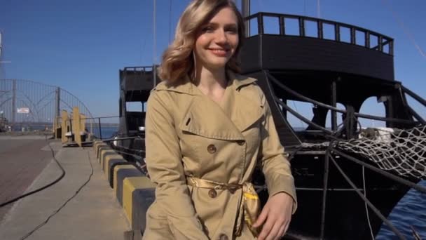 Привлекательная молодая блондинка в окопе с винтажным чехлом сидит на причале Jacht Pier and Points in the Sea. Темный корабль на заднем плане — стоковое видео