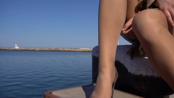Attraktive junge blonde Frau im Trench mit Vintage-Koffer sitzt auf der Seebrücke. Neigung nach oben. — Stockvideo