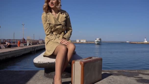 Atractiva joven rubia en trinchera con maleta vintage está sentada en Bollard en el muelle Jacht — Vídeo de stock