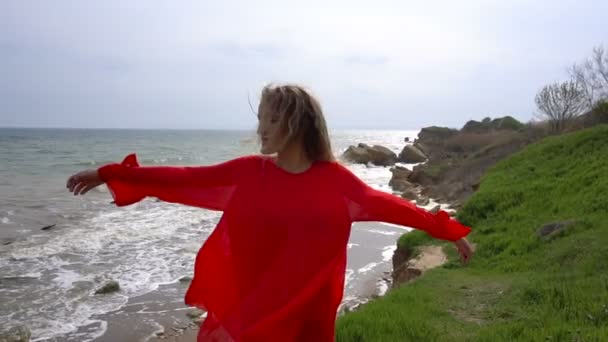 Счастливая молодая женщина в красном платье кружит и веселится на утесе морского побережья — стоковое видео