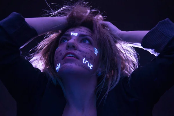 Lány szintetizált hajjal drámai lila fényben, különböző nyelvi szavakkal az arcán. Gyönyörű modell UV fluoreszkáló neon kreatív make-up. arccal közelről — Stock Fotó