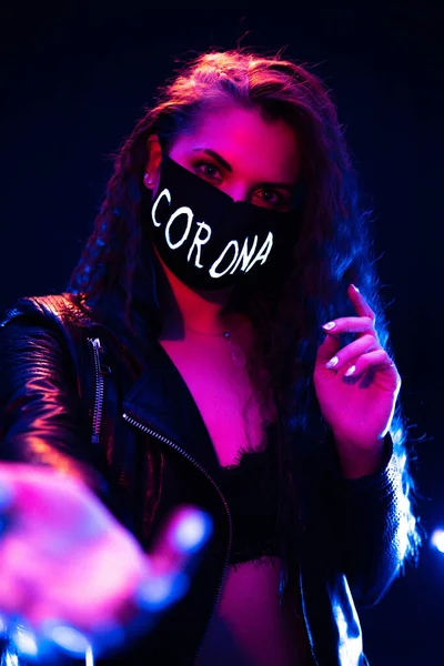 Retrato de una atractiva chica atractiva usando una máscara protectora negra con el cartel de Corona en la cara en luces de neón con fondo de niebla negra — Foto de Stock