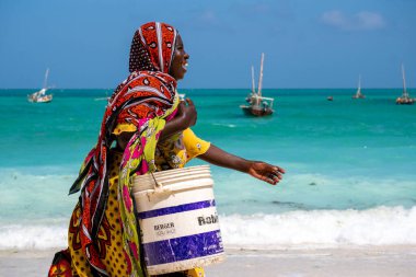 NUNGWI, ZANZIBAR - 20 HAZİRAN 2019: Hint Okyanus Sahili kıyısında, Tanzanya 'nın Zanzibar adasındaki Balık Nungwi köyü için bir kova ile yürüyen Parlak Kumaşlı Kadın