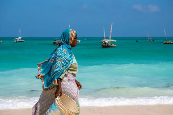 NUNGWI, ZANZIBAR - JAN 20, 2019: místní žena v Bright Cloth kráčí podél Indického oceánu Beach s kyblíkem pro ryby vesnice Nungwi, Zanzibar ostrov, Tanzanie — Stock fotografie