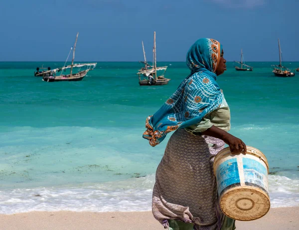NUNGWI, ZANZIBAR - JAN 20, 2019: μια ντόπια γυναίκα με φωτεινό πανί που περπατάει δίπλα στην παραλία του Ινδικού Ωκεανού με ένα κουβά για το χωριό Fish Nungwi, νησί Zanzibar, Τανζανία — Φωτογραφία Αρχείου