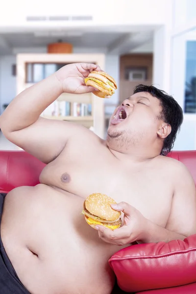 Fat man eating burgers on the sofa — Stock fotografie