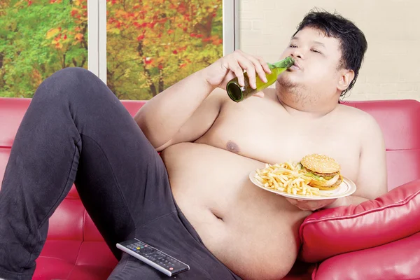 Overweight man drinking and eating on couch