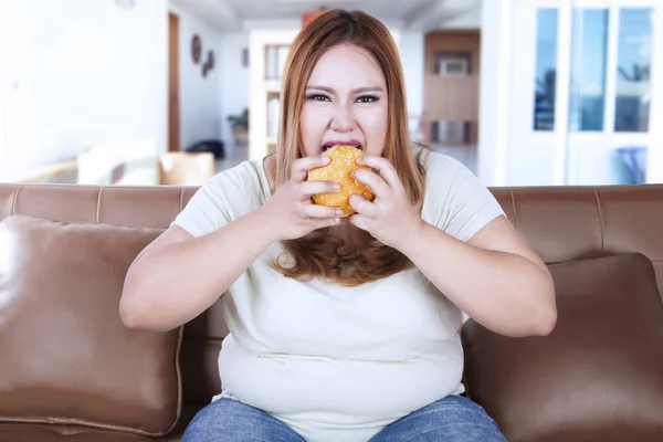 Mujer obesa comiendo hamburguesa en el sofá —  Fotos de Stock