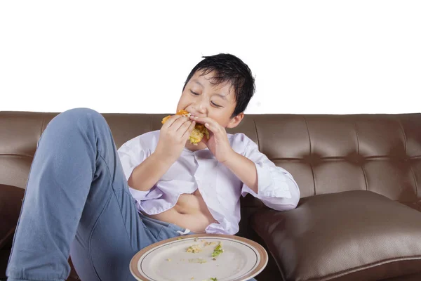 Little boy eats cheeseburger on couch — Stock Photo, Image