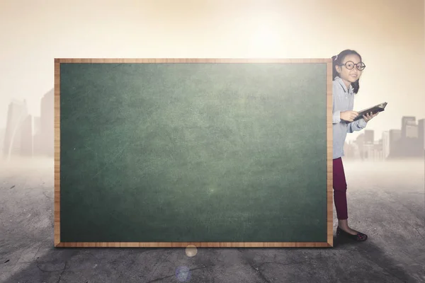 Little girl with book behind a chalkboard — Stock Photo, Image