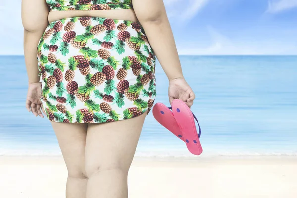 Mujer con sobrepeso sostiene sandalias en la playa — Foto de Stock