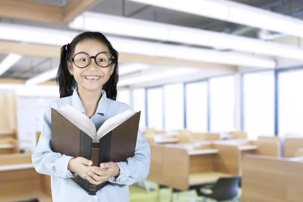 Colegiala sonriendo a la cámara con libro en el aula — Foto de Stock